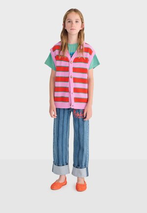 Tyttö, jolla on päällään vaaleanpunainen ja punainen raidallinen neulottu liivi vihreän t-paidan päällä, siniset käännetyt farkut, joissa on tekstiä, ja oranssit liukusäätösandaalit, seisoo yksinkertaisen taustan edessä.