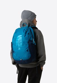 Kék nejlon hátizsák párnázott hátrésszel, hálós oldalzsebekkel, gumis rögzítéssel és nagy logóval. Szürke kapucnis pulóverrel viselve.