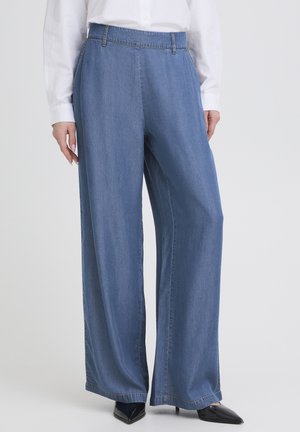 Naine, kes kannab laiade säärega siniseid teksapükse, mustade teravate ninaosadega kontsakingi ja valget nööbitavat särki tavalise taustaga.