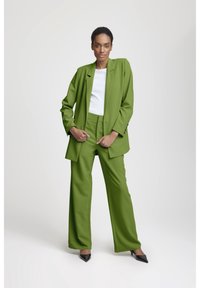 Kvinna som står självsäkert i en grön oversized kostym, vit topp och svarta spetsiga klackskor mot en ljus och enkel bakgrund.