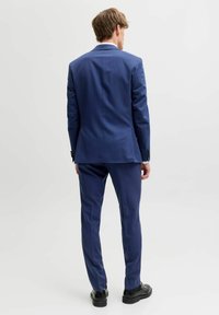 Vīrietis stāv ar muguru pret skatītāju, ģērbies pieguļošā zilā uzvalkā un melnās formālās kurpēs, uz vienkārša gaiša fona.