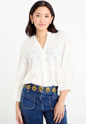 Femme portant un chemisier blanc en broderie anglaise à manches trois quarts et un jean bleu avec une ceinture à médaillon doré, debout devant un fond blanc uni.