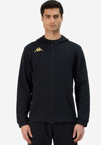 Felpa nera con zip realizzata in tessuto morbido, caratterizzata da un logo dorato sulla spalla sinistra e due tasche frontali. Progettata con una vestibilità comoda.