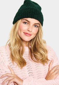 Green knitted beanie with a soft texture, worn by a person with long blonde hair, paired with a light pink, eyelet-patterned sweater.