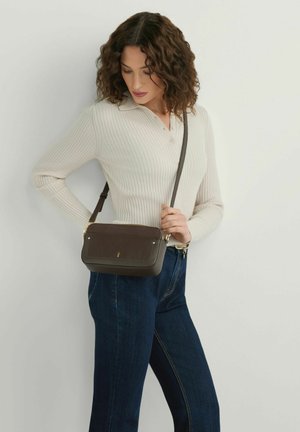 Kobieta o kręconych włosach, ubrana w kremowy prążkowany sweter, ciemnoniebieskie dżinsy i ciemnobrązową torebkę na ramię, stojąca na białym tle.