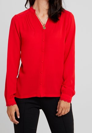 Kobieta ubraną w jasnoczerwoną, zapinaną na guziki koszulę z długim rękawem zakończoną koronkowymi mankietami oraz złotym wisiorkiem na szyi, w połączeniu z czarnymi spodniami.