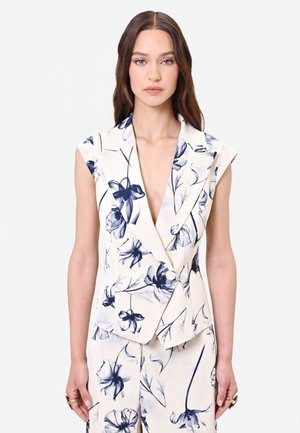 Femme aux longs cheveux bruns portant un blazer blanc sans manches et un pantalon assorti avec des motifs floraux bleus sur un fond clair uni.