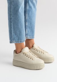 Kremowe skórzane sneakersy z futrzanym kołnierzem, o zaokrąglonym nosku i grubej, żłobkowanej podeszwie. Posiadają złote oczka i beżowe sznurowadła.