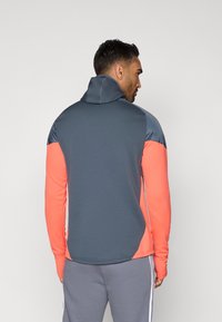 Herre træningsjakke med en grå krop og koralfarvede ærmer samt skulderdetaljer. Lavet af et glat og fleksibelt materiale med høj krave.