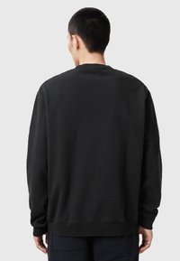 Personne vue de dos portant un sweat-shirt noir uni et un pantalon sombre devant un arrière-plan gris clair uni.