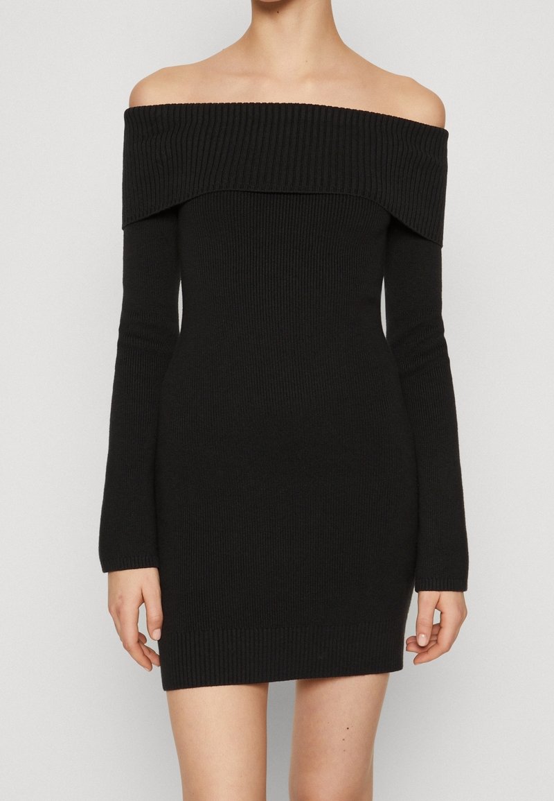 Femme portant une mini-robe noire en maille côtelée, épaules dénudées et manches longues, sur un fond clair uni.