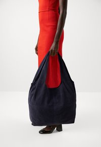 Navy suede bag with a large, open-top design and dual side pockets, held by a person wearing a fitted red dress and black heels.