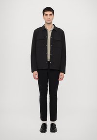 Jachetă neagră cu buzunare mari pe piept, combinată cu un top bej deschis și pantaloni negri ajustați. Modelul poartă pantofi negri.