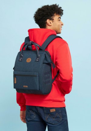 Dunkelblauer Rucksack aus strukturiertem Stoff, mit zwei Ledergriffen, einer vorderen Reißverschlusstasche und verstellbaren Schulterriemen. Akzentuiert mit einem braunen Logo.
