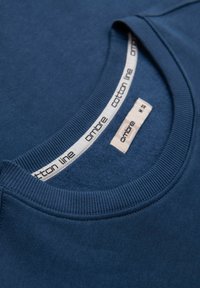 Gros plan d'un sweat-shirt bleu à col rond montrant un col côtelé et une étiquette de taille blanche indiquant "ombre M 38" à l'intérieur de l'encolure.