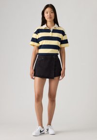 Νεαρή γυναίκα που φοράει κίτρινη και ναυτική ριγέ πόλο μπλούζα, μαύρη ασύμμετρη μίνι φούστα και λευκά αθλητικά Nike, στέκεται σε ουδέτερο φόντο.