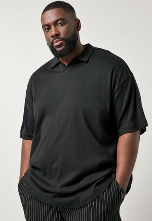 Brodaty mężczyzna w luźnej czarnej koszulce polo i czarnych spodniach w prążki, stojący z rękami w kieszeniach na jednolitym jasnym tle.