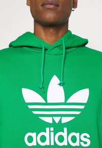 Zelená mikina s kapucí z měkkého materiálu s velkým bílým logem Adidas se třemi pruhy a charakteristickým motivem listu.