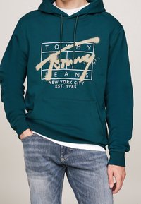 Hombre que lleva una sudadera color verde azulado con el texto "Tommy Jeans New York City Est. 1985" y un logo en estilo pintura, combinada con unos vaqueros azules desgastados.