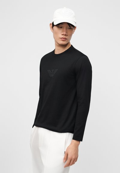 Νέος άνδρας που φοράει μαύρο μακρυμάνικο πουκάμισο με διακριτικό λογότυπο και λευκό παντελόνι, συνδυασμένο με άσπρο καπέλο, μπροστά σε απλό φόντο.