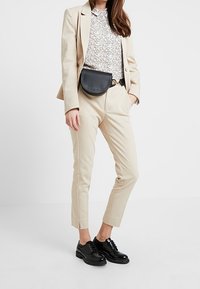 Femme portant un costume beige avec une blouse à motifs, un sac ceinture noir et des chaussures oxford brillantes noires, se tenant avec une main dans la poche.