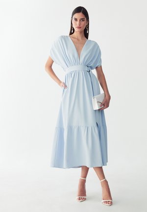 Woman in light blue midi dress with deep V-neck, puffed sleeves, cinched waist, holding white clutch, wearing white strappy heels and drop earrings.