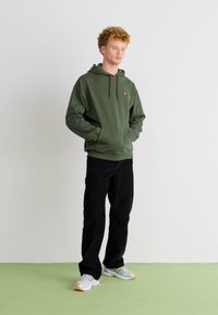 Sudadera de color verde oliva con bolsillo frontal y cordones, combinada con pantalones negros. El modelo lleva zapatillas gris claro. Fondo minimalista.