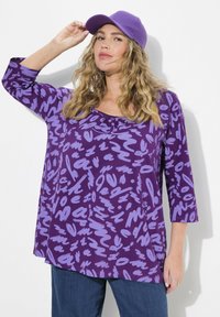 Lila bedruckte Bluse mit dreiviertelärmeln, die abstrakte, helllila Muster aufweist. Model trägt eine passende lila Mütze.