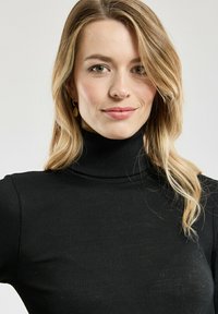 Junge Frau mit blonden, gewellten Haaren, die einen schwarzen Rollkragenpullover und goldene Ohrringe trägt, lächelt sanft vor einem neutralen Hintergrund.