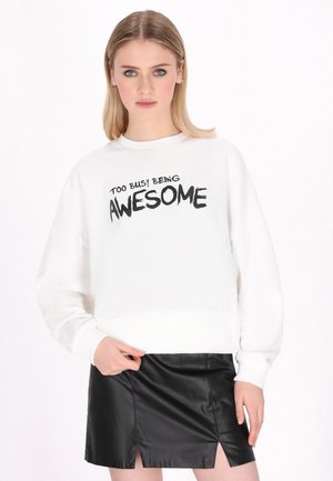 Młoda kobieta o blond włosach, ubrana w biały sweter z napisem „Too Busy Being Awesome” oraz krótką czarną skórzaną spódniczkę.