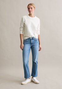 Lichtgewicht witte trui met een ronde hals, gecombineerd met lichtblauwe jeans met rechte pijpen en klassieke witte sneakers. Achtergrond van een vlakke ondergrond.