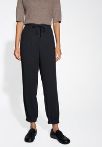 Pantaloni neri a tapered con vita regolabile, dotati di due tasche frontali e polsini elasticizzati. Indossati con scarpe nere. La texture appare liscia.