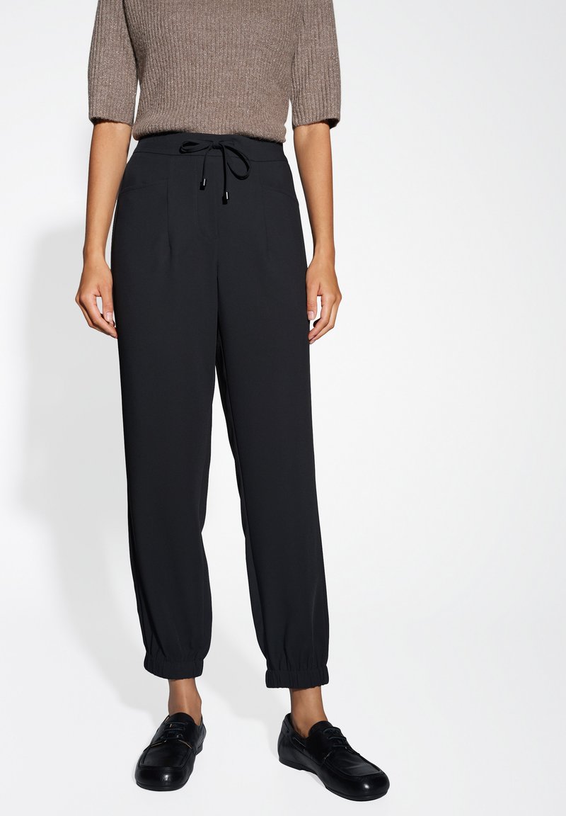 Pantaloni neri a tapered con vita regolabile, dotati di due tasche frontali e polsini elasticizzati. Indossati con scarpe nere. La texture appare liscia.