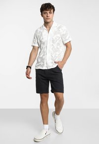 Witte overhemd met korte mouwen en een geometrisch patroon, gecombineerd met donkere shorts en witte instap schoenen. Casual ontwerp en pasvorm.
