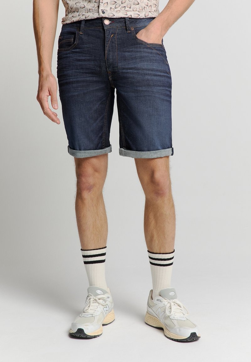 Donkerdenim korte broek met opgerolde boorden, met een vijf-pocket ontwerp en contrasterende stiksels. Gecombineerd met witte sneakers en gestreepte sokken.