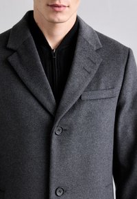 Cappotto di lana grigia con un design strutturato, caratterizzato da un rever a preferenza, tre bottoni e una tasca sul petto. Lo strato interno ha una texture a coste nera.