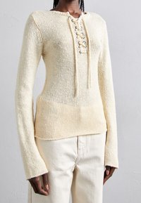 Pull en tricot de couleur crème avec encolure ronde, devant à lacets et manches longues. La texture est lâche et texturée, avec une finition mélangée.