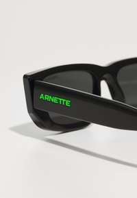 Ochelari de soare negri cu un cadru grozav, având o finisare lucioasă. "ARNETTE" în litere verzi strălucitoare pe lateral, lentile fumurii negre.