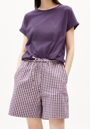 Frau trägt ein lila T-Shirt mit hochgekrempelten Ärmeln, das in hoch taillierte, lila-weiß karierte Shorts mit Tunnelzug gesteckt ist, Hände in den Taschen.