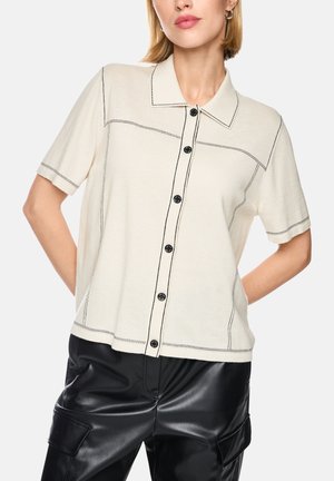 Femme portant une chemise crème à manches courtes boutonnée avec des surpiqûres noires en contraste et un pantalon en cuir noir avec poches.
