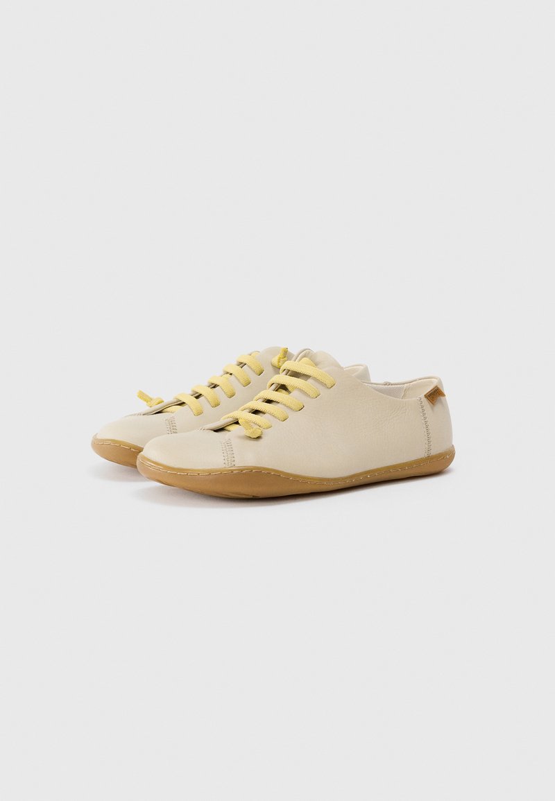 Chaussures en cuir beige avec des lacets jaunes et des semelles en caoutchouc, présentant des détails de couture et une forme arrondie. Design minimaliste, style à tige basse.