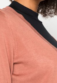 Nära bild av en person som bär en svart höghalshalsad topp under en dammig rosa cardigan, med lockigt hår synligt mot en neutral bakgrund.