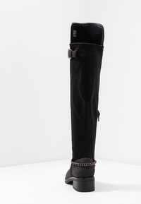 Botas negras hasta la rodilla que presentan un material suave, un acento de cadena texturizada en el tacón y una forma estrecha. Detalle de cremallera lateral visible.