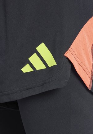 Zwarte sportshirt met oranje mesh accenten; heeft een opvallend groen drie-strepenlogo op de onderste zoom. De textuur lijkt glad te zijn.