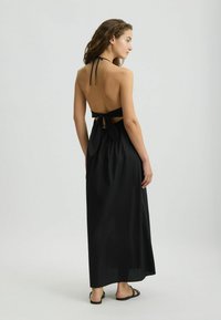 Czarna maxi sukienka z halter neck, odkrytymi plecami i wiązaniem w talii. Gładki materiał opada na ziemię, dopełniony prostymi sandałami.