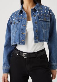 Lyhyt farkkutakki, sininen, hopeanvärisillä niiteillä hartioissa, napit edessä, kaksi taskua. Yhdistetty valkoisen paidan ja mustan vyön kanssa mustien farkkujen päällä.