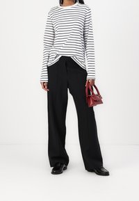Donna che indossa una camicia bianca a maniche lunghe con strisce nere, pantaloni neri a gamba larga, mocassini neri, e tiene in mano una piccola borsa rossa.