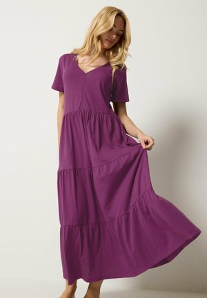 Femme en robe maxi violette à étages avec des manches courtes, tenant la jupe tout en se tenant contre un fond clair uni.