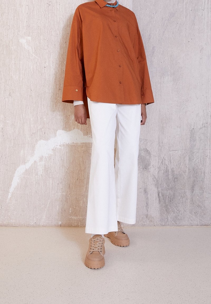 Personne portant une chemise ample orange brûlé à boutons, pantalon blanc à jambes larges, baskets beige épaisses, debout contre un mur texturé.