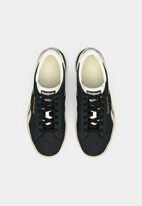 Crne suede tenisice s bijelim naglascima, okrugli vrh, crne vezice i teksturirana gumena potplata. Sadrži "Reebok" oznaku sa strane.